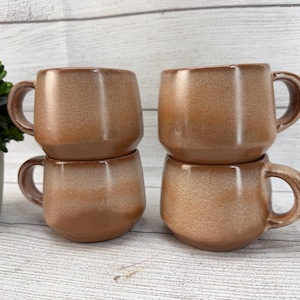 May include: Four brown ceramic mugs with rounded handles are stacked in pairs. The mugs have a speckled, glazed finish with a gradient from light to dark brown. The mugs are on a light wood-grain surface.