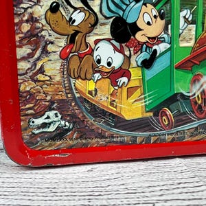 May include: Red metal lunchbox featuring Mickey Mouse, Donald Duck, and Goofy riding a train. The train is green and yellow with a red smokestack. The lunchbox has the text "MCM LXXX Walt Disney Productions" and "ALADDIN INDUSTRIES, INCORPORATED, NASHVILLE, TN."