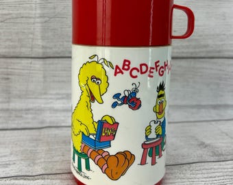 May include: A red and white Sesame Street thermos with a yellow Big Bird character reading a book that says "Lunch".  A blue bee with a red apple and a red-haired character with a blue shirt and a white beard are also on the thermos. The letters A, B, C, D, E, F, G, and H are printed on the thermos.