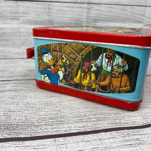 May include: A vintage metal lunchbox featuring a cartoon image of Donald Duck and three pirates in a jail cell. The lunchbox is red with a blue and white interior.