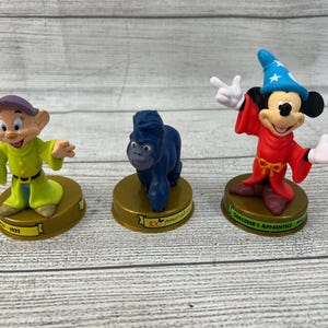May include: Three Disney character figurines: Dopey in a green outfit, a blue gorilla, and Mickey Mouse in a red robe and blue wizard hat. Each figure is on a gold-colored base. The Mickey Mouse figure is labeled "Sorcerer's Apprentice".