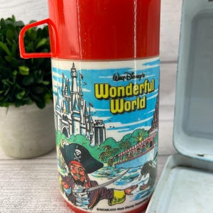 Op de afbeelding: Vintage rode en witte Disney's Wonderful World thermoskan met een rode handgreep. De thermoskan heeft een piratenillustratie en een kasteelsc&egrave;ne. Een lichtblauwe lunchbox staat op de achtergrond.