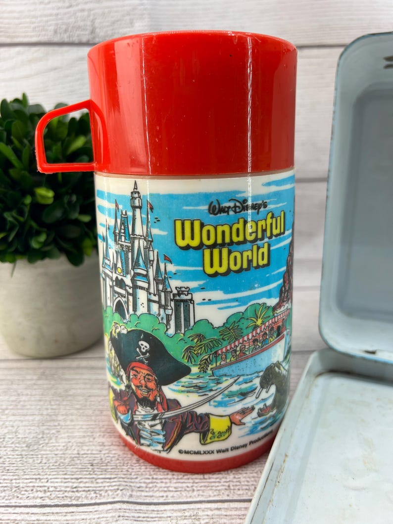Op de afbeelding: Vintage rode en witte Disney's Wonderful World thermoskan met een rode handgreep. De thermoskan heeft een piratenillustratie en een kasteelsc&egrave;ne. Een lichtblauwe lunchbox staat op de achtergrond.