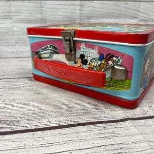 May include: Vintage red and blue metal lunchbox featuring a scene of Mickey Mouse and friends at Disney World. The lunchbox has a red handle and a metal latch.