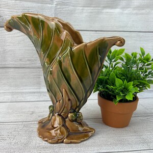 May include: A ceramic vase with a leafy design in shades of green and brown. The vase is shaped like a leaf and has a wide opening at the top.