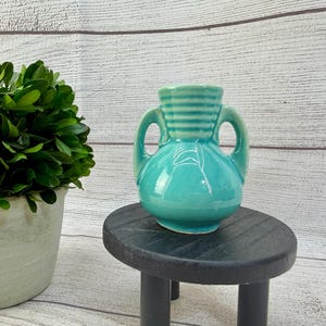May include: A small, turquoise ceramic vase featuring a rounded body, two side handles, and a ribbed neck design. The piece has a glossy finish and a smooth, curved silhouette, standing as a decorative vessel with a classic, symmetrical form.