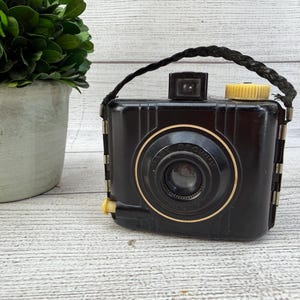 May include: A vintage black Baby Brownie camera featuring a rectangular body with vertical ribbed detailing, a central circular lens housing with gold-tone trim, a yellow winding knob, and a braided black carrying strap attached to the sides.