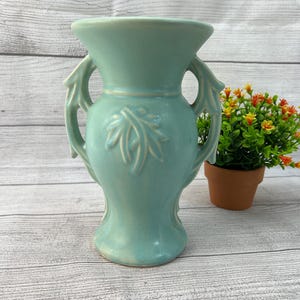 May include: A light blue ceramic vase with two handles and a leaf design. The vase is on a white wooden surface.