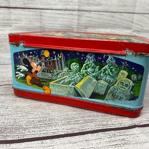 May include: Vintage red and blue metal lunchbox featuring Mickey Mouse running away from a haunted house with ghosts and tombstones. The lunchbox has a 'RIP' inscription on one of the tombstones.