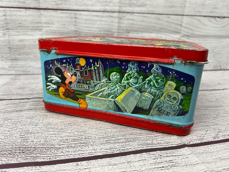 May include: Vintage red and blue metal lunchbox featuring Mickey Mouse running away from a haunted house with ghosts and tombstones. The lunchbox has a 'RIP' inscription on one of the tombstones.