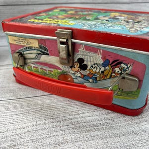 Op de afbeelding: Vintage metalen lunchbox met een rood frame en handvat. De zijkanten tonen Disney-personages Mickey Mouse, Donald Duck, Goofy en anderen in een ruimtethema-ontwerp. De bovenkant heeft een kleurrijke illustratie van een themapark.