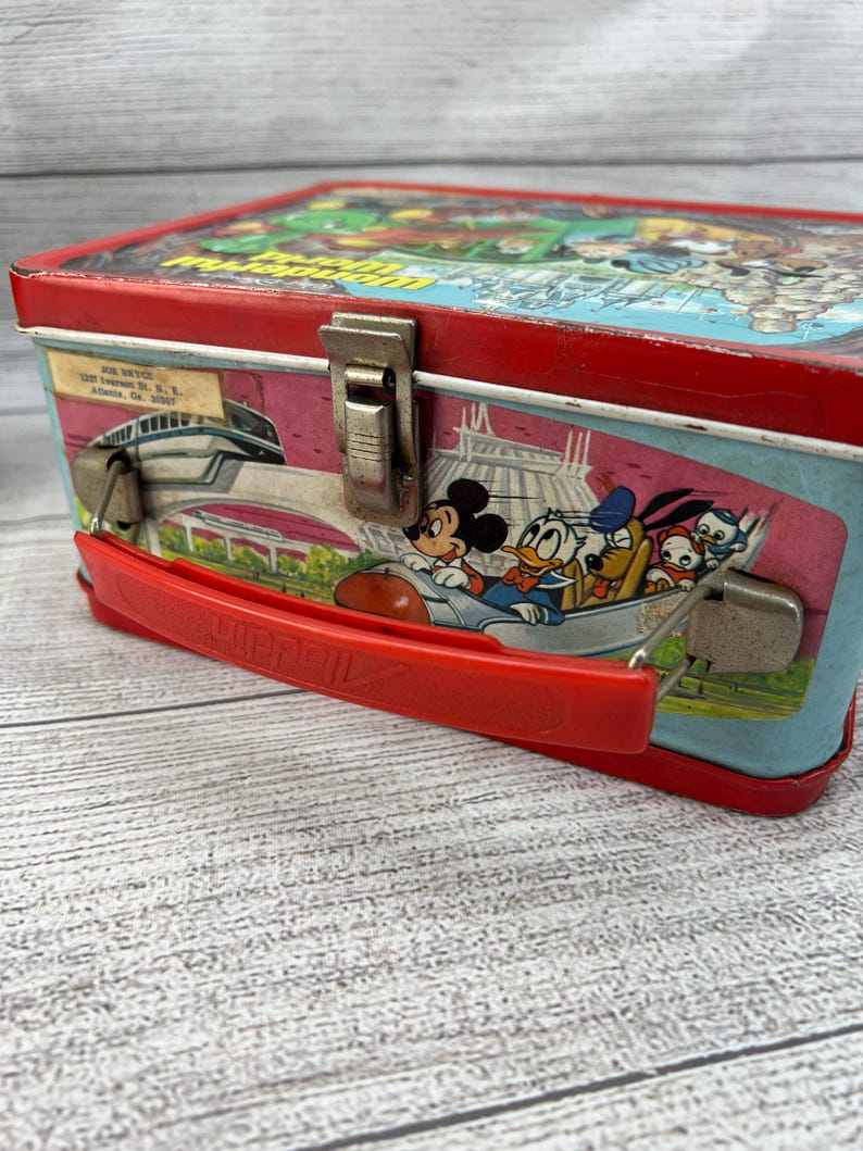 Op de afbeelding: Vintage metalen lunchbox met een rood frame en handvat. De zijkanten tonen Disney-personages Mickey Mouse, Donald Duck, Goofy en anderen in een ruimtethema-ontwerp. De bovenkant heeft een kleurrijke illustratie van een themapark.