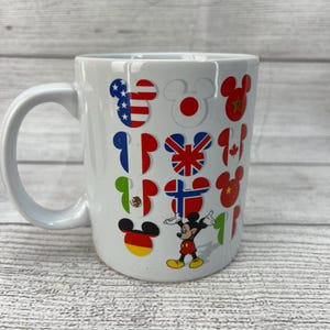 Puede incluir: Taza de caf&eacute; de cer&aacute;mica blanca con un dise&ntilde;o de Mickey Mouse. La taza presenta a Mickey Mouse en el centro con los brazos extendidos. Alrededor de Mickey Mouse hay orejas de Mickey Mouse con banderas de diferentes pa&iacute;ses dentro de cada oreja.