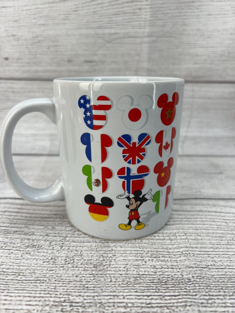 Puede incluir: Taza de caf&eacute; de cer&aacute;mica blanca con un dise&ntilde;o de Mickey Mouse. La taza presenta a Mickey Mouse en el centro con los brazos extendidos. Alrededor de Mickey Mouse hay orejas de Mickey Mouse con banderas de diferentes pa&iacute;ses dentro de cada oreja.