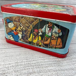 Op de afbeelding: Vintage metalen lunchbox met een rode boven- en onderkant en een lichtblauwe behuizing. De lunchbox bevat cartoon illustraties van Donald Duck en piraten. De lunchbox is rechthoekig en heeft een handvat.