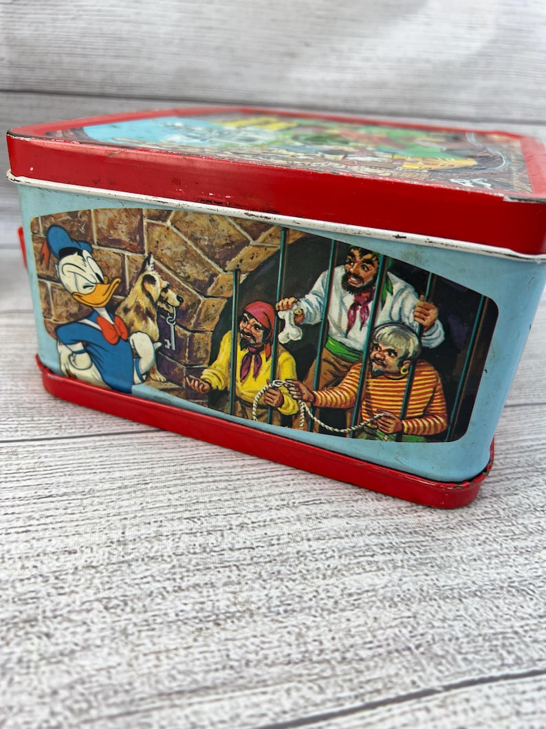 Op de afbeelding: Vintage metalen lunchbox met een rode boven- en onderkant en een lichtblauwe behuizing. De lunchbox bevat cartoon illustraties van Donald Duck en piraten. De lunchbox is rechthoekig en heeft een handvat.