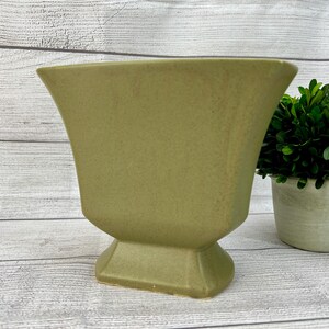May include: A green ceramic vase with a square base and a wide, flared opening. The vase has a textured surface and a slightly distressed look.