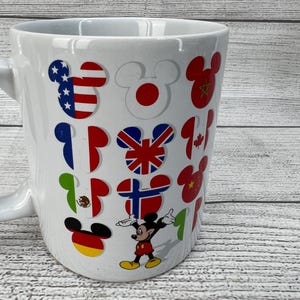 Puede incluir: Taza de cer&aacute;mica blanca con Mickey Mouse y banderas de diferentes pa&iacute;ses del mundo. Las banderas incluyen Estados Unidos, Jap&oacute;n, China, Reino Unido, Canad&aacute;, M&eacute;xico, Alemania y Finlandia.