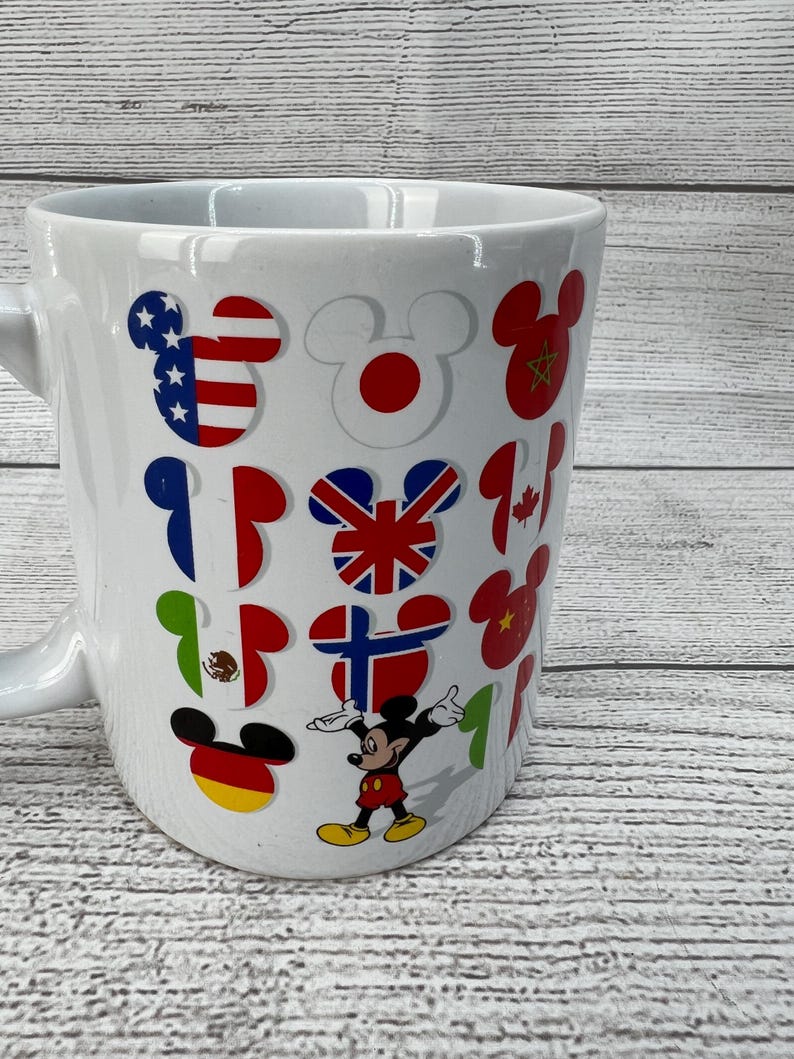 Puede incluir: Taza de cer&aacute;mica blanca con Mickey Mouse y banderas de diferentes pa&iacute;ses del mundo. Las banderas incluyen Estados Unidos, Jap&oacute;n, China, Reino Unido, Canad&aacute;, M&eacute;xico, Alemania y Finlandia.