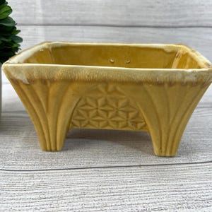 May include: A square, yellow ceramic planter with a textured geometric pattern on the sides and a slightly lighter rim. The planter is elevated on three small feet. The planter is suitable for small plants.
