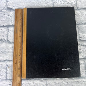 May include: A black hardcover notebook with a brown spine and a gold-colored signature on the front cover that reads "Ellen Feldman".