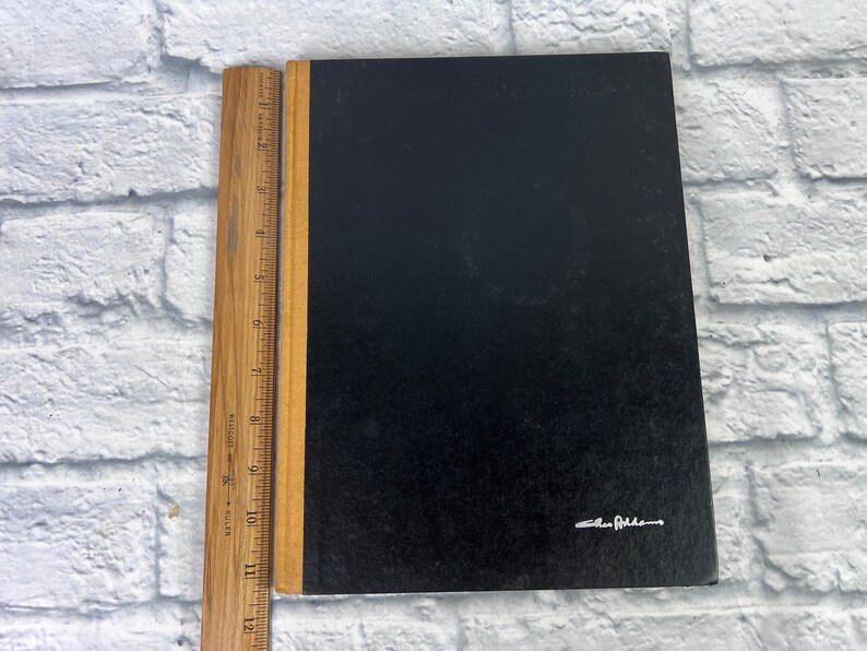May include: A black hardcover notebook with a brown spine and a gold-colored signature on the front cover that reads "Ellen Feldman".