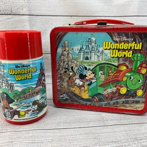 Op de afbeelding: Vintage rode metalen lunchbox en bijpassende thermoskan met Disney's Wonderful World. De lunchbox heeft een rode handgreep en een kleurrijke illustratie van Mickey Mouse, Pluto en Donald Duck op een trein. De thermoskan is rood met een blauw-witte illustratie.