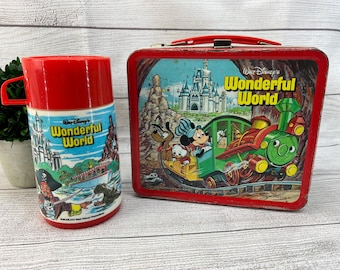 May include: Vintage red metal lunchbox and matching thermos featuring Disney's Wonderful World. The lunchbox has a red handle and a colorful illustration of Mickey Mouse, Pluto, and Donald Duck on a train. The thermos is red with a blue and white illustration.