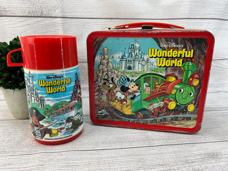 Op de afbeelding: Vintage rode metalen lunchbox en bijpassende thermoskan met Disney's Wonderful World. De lunchbox heeft een rode handgreep en een kleurrijke illustratie van Mickey Mouse, Pluto en Donald Duck op een trein. De thermoskan is rood met een blauw-witte illustratie.
