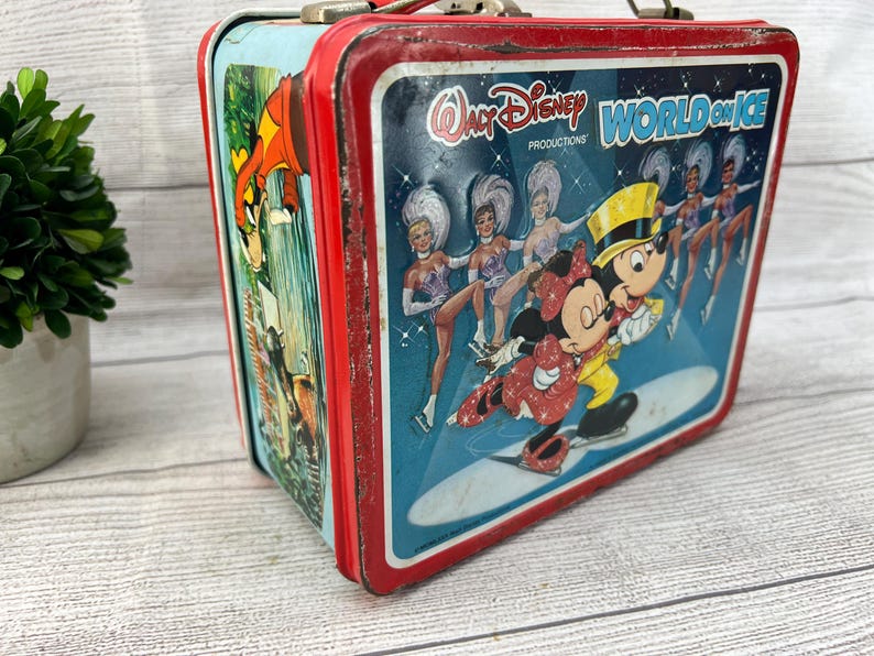 Op de afbeelding: Vintage metalen lunchbox met een rood frame en een blauwe achtergrond. De lunchbox toont Mickey en Minnie Mouse schaatsen, met de tekst "Walt Disney World on Ice". Het zijpaneel heeft een cartoon illustratie.