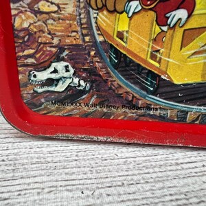 Op de afbeelding: Vintage rode Aladdin lunchbox met een cartoon treinsc&egrave;ne en een dinosaurus schedel. De lunchbox heeft een rode rand en vertoont tekenen van slijtage. De tekst "&copy; MCMLXXX Walt Disney Productions" is inbegrepen.