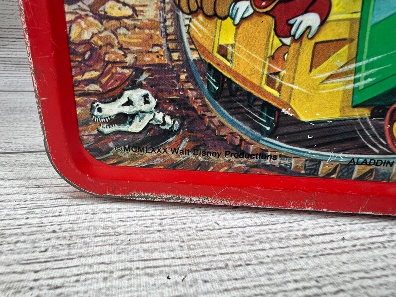 Op de afbeelding: Vintage rode Aladdin lunchbox met een cartoon treinsc&egrave;ne en een dinosaurus schedel. De lunchbox heeft een rode rand en vertoont tekenen van slijtage. De tekst "&copy; MCMLXXX Walt Disney Productions" is inbegrepen.