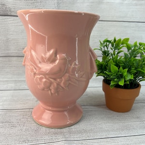May include: A pink ceramic vase with a bird and floral design. The vase has a wide mouth and a narrow base.