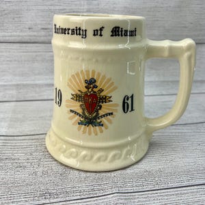 May include: A white ceramic beer stein with a black inscription that reads "University of Miami" and a crest with the letters "TKA" and the numbers "19" and "61".