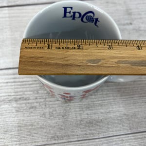 Puede incluir: Taza de cer&aacute;mica blanca con un logotipo de Epcot azul y blanco y un dise&ntilde;o rojo y blanco. La taza est&aacute; sobre una superficie de madera.