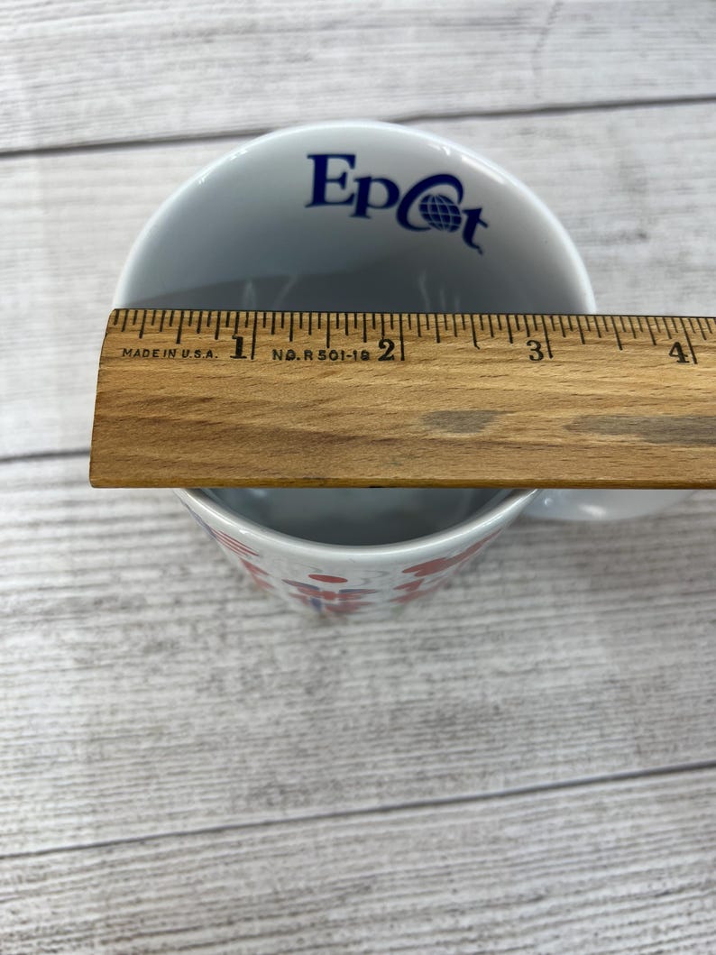 Puede incluir: Taza de cer&aacute;mica blanca con un logotipo de Epcot azul y blanco y un dise&ntilde;o rojo y blanco. La taza est&aacute; sobre una superficie de madera.