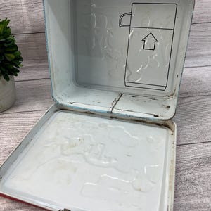 Op de afbeelding: Vintage metalen lunchbox met een rode handgreep en rand, en een witte binnenkant. De binnenkant heeft een reli&euml;f ontwerp en een zwarte omtrek van een thermoskan met een pijl. De lunchbox is open.