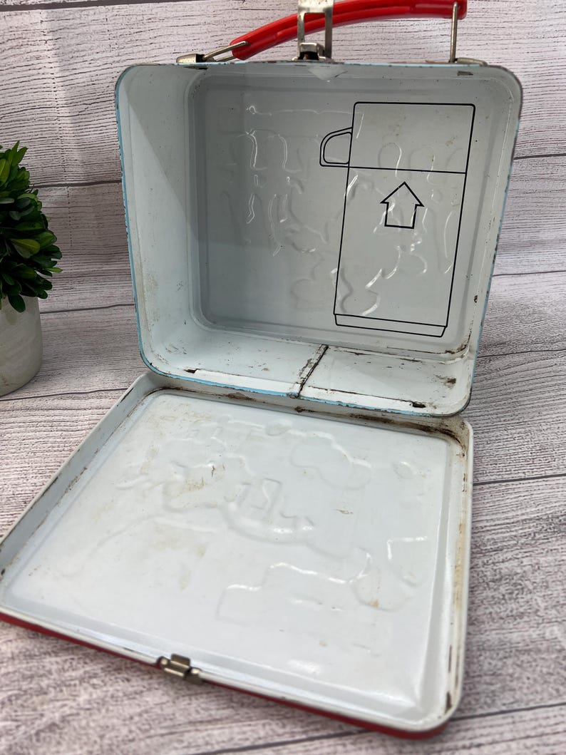 Op de afbeelding: Vintage metalen lunchbox met een rode handgreep en rand, en een witte binnenkant. De binnenkant heeft een reli&euml;f ontwerp en een zwarte omtrek van een thermoskan met een pijl. De lunchbox is open.