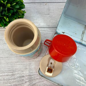 Op de afbeelding: Vintage Aladdin thermoskan met een rode beker en een lichtblauwe lunchbox. De thermoskan heeft een beige behuizing met een kleurrijk patroon. De beker en het deksel zijn los van de thermoskan. De lunchbox is open.