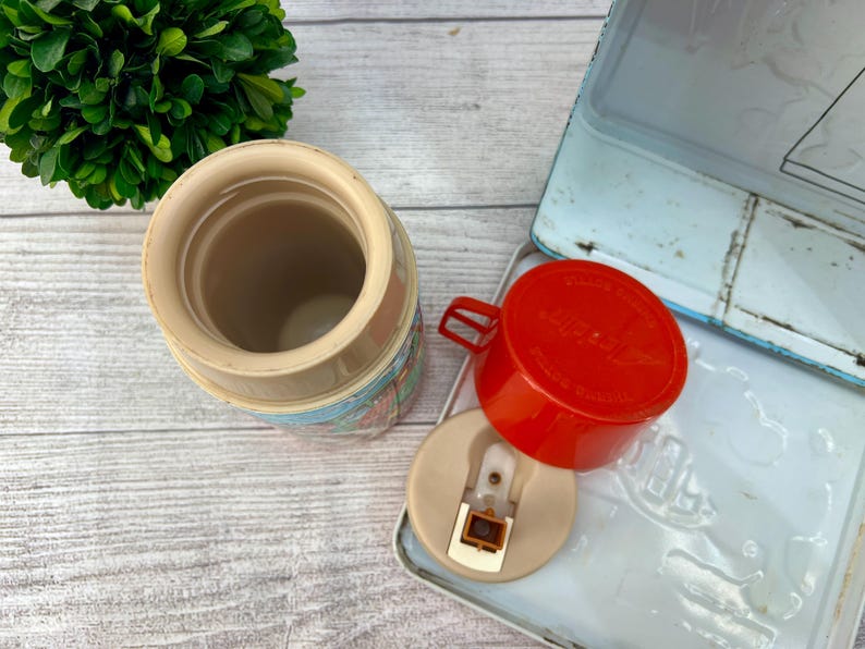 Op de afbeelding: Vintage Aladdin thermoskan met een rode beker en een lichtblauwe lunchbox. De thermoskan heeft een beige behuizing met een kleurrijk patroon. De beker en het deksel zijn los van de thermoskan. De lunchbox is open.