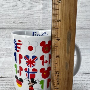 Puede incluir: Taza de cer&aacute;mica blanca con un dise&ntilde;o de Mickey Mouse con banderas de diferentes pa&iacute;ses. La taza tiene un asa y dice "Epcot" en el lateral.