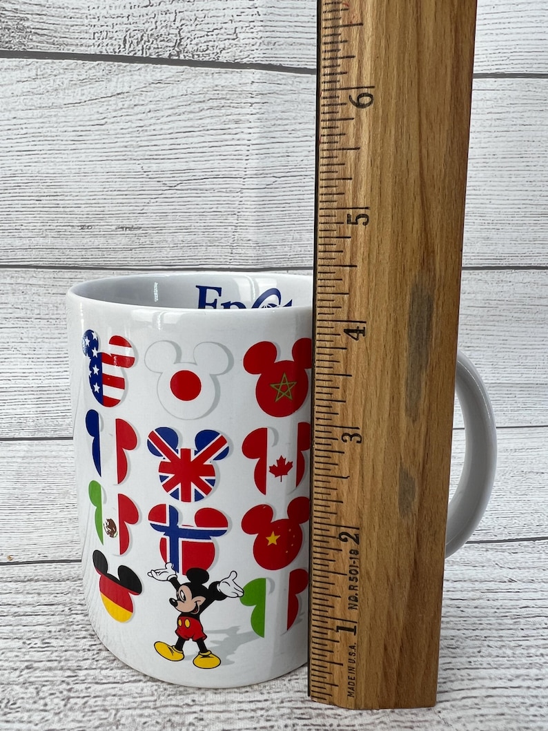 Puede incluir: Taza de cer&aacute;mica blanca con un dise&ntilde;o de Mickey Mouse con banderas de diferentes pa&iacute;ses. La taza tiene un asa y dice "Epcot" en el lateral.