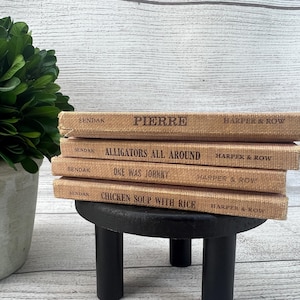 May include: A stack of four vintage children's books with tan covers and black text. The titles include "Pierre," "Alligators All Around," "One Was Johnny," and "Chicken Soup with Rice." The books are stacked on a small, black, three-legged table.