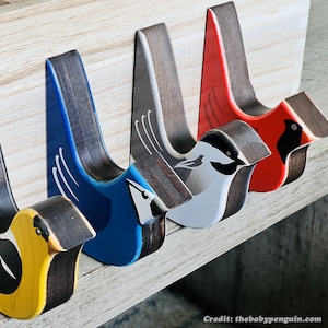 May include: Five wooden bird-shaped hooks painted in different colors, including yellow, blue, white, red, and black. The hooks are mounted on a light wood surface.