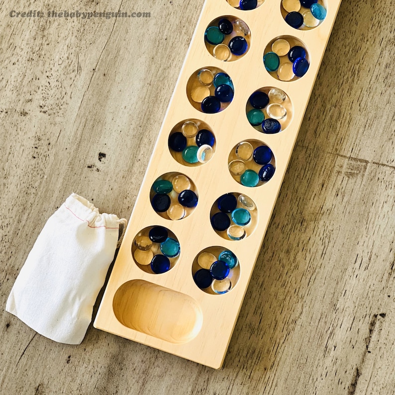 May include: A wooden game board with a row of 14 circular depressions, each containing blue and clear glass marbles. The board is made of light wood and has a natural finish.