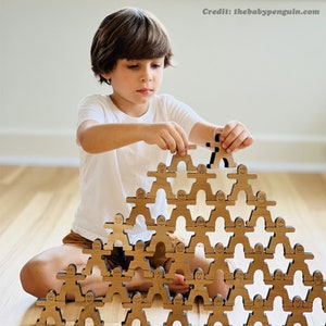 May include: A child sits on the floor building a tower with wooden stacking people. The tower is made of multiple layers of wooden people with different facial expressions. The child is carefully placing a wooden person on top of the tower.