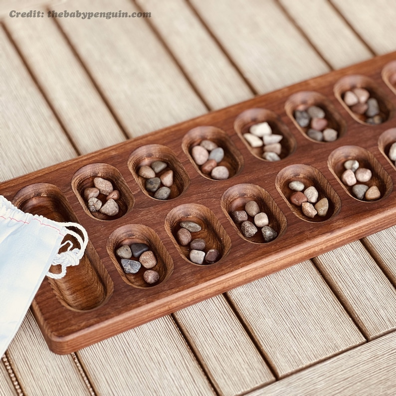 May include: A wooden game board with 14 compartments, each containing small stones. The board is made of dark wood and has a smooth, polished finish. The stones are a variety of colors and sizes.