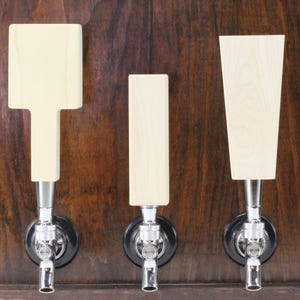 May include: Three wooden beer tap handles in different shapes and sizes. The handles are made of light wood and have a natural finish. They are mounted on a dark wood background.