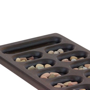 May include: A wooden game board with 12 oval-shaped depressions, each filled with small, colorful stones. The board is dark brown and has a smooth, polished finish.