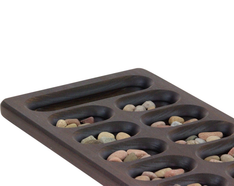 May include: A wooden game board with 12 oval-shaped depressions, each filled with small, colorful stones. The board is dark brown and has a smooth, polished finish.